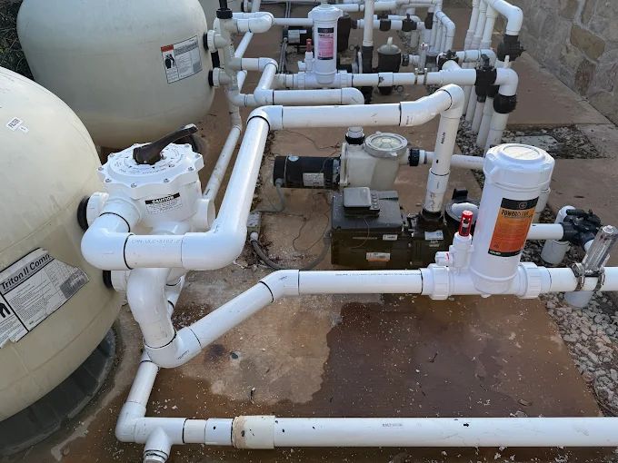 Pool filtration system with white PVC pipes, a pump, and a filter in a backyard setting; multiple valves and gauges visible on concrete patio.
